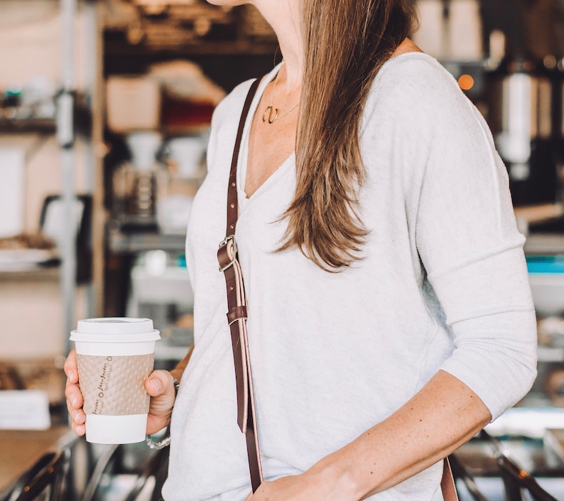 May include: A woman wearing a white long-sleeved shirt with a brown leather strap. She is holding a white to-go coffee cup with brown text that says "Brown Paper Bag".