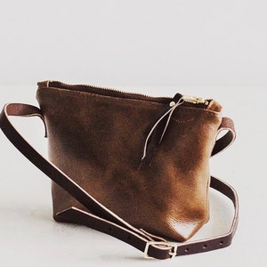 May include: A brown leather crossbody bag with a zipper closure and a long adjustable strap.
