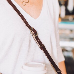 May include: A person wearing a white v-neck shirt with a brown leather strap and a gold necklace. The strap has a gold buckle. The person is holding a white to-go coffee cup.