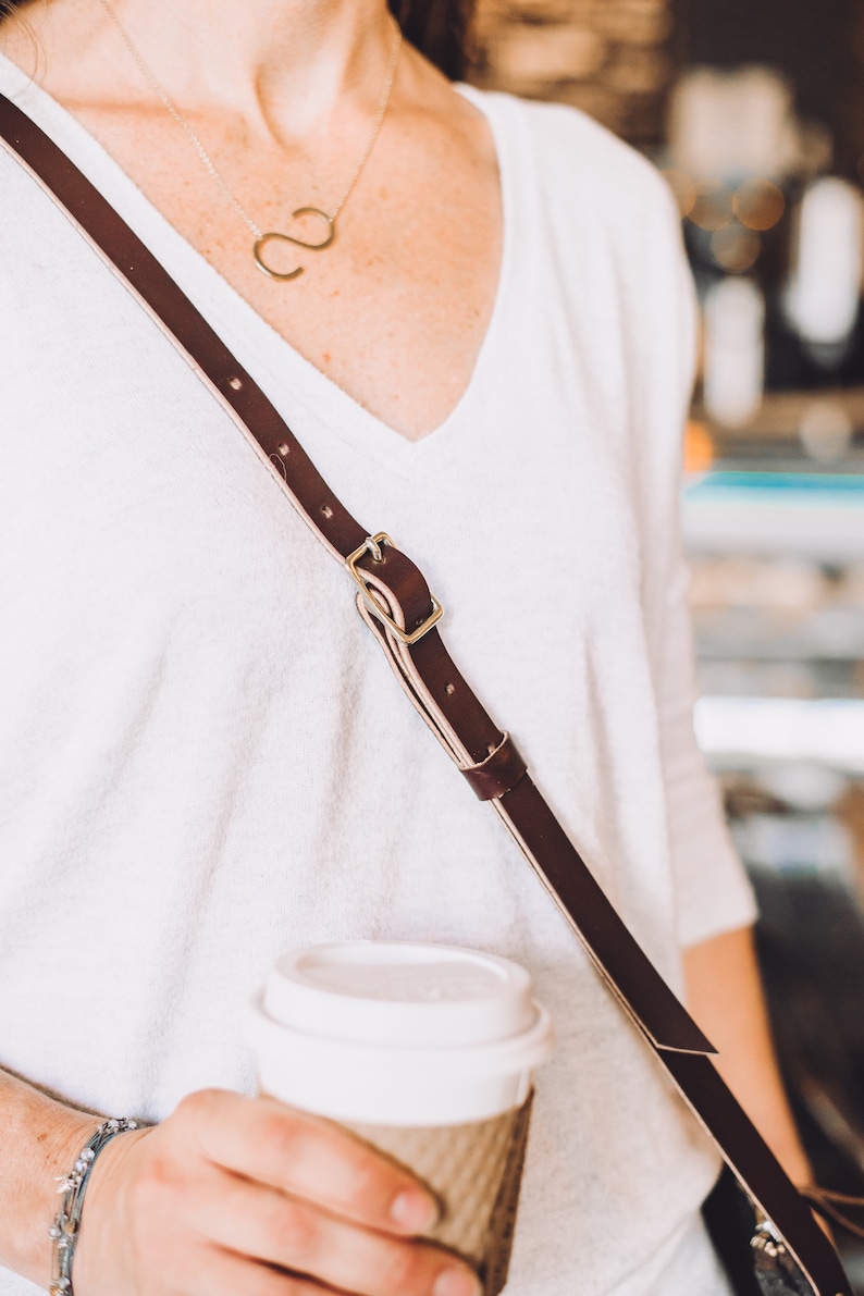 May include: A person wearing a white v-neck shirt with a brown leather strap and a gold necklace. The strap has a gold buckle. The person is holding a white to-go coffee cup.