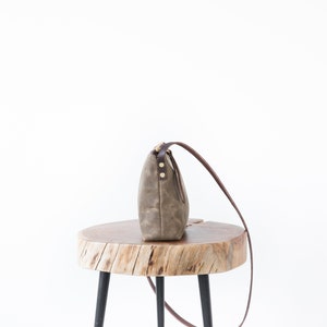 May include: A brown canvas crossbody bag with a brown leather strap and gold accents sits on a wooden stool with black legs.
