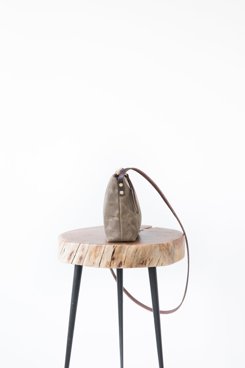 May include: A brown canvas crossbody bag with a brown leather strap and gold accents sits on a wooden stool with black legs.