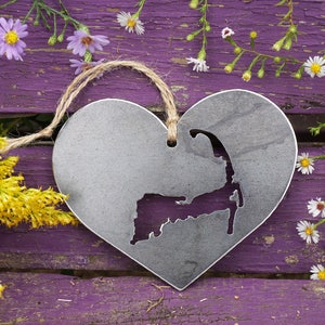 May include: A metal heart-shaped ornament with a cutout of the shape of Cape Cod. The ornament is hanging from a twine string and is on a purple wooden surface with white flowers.