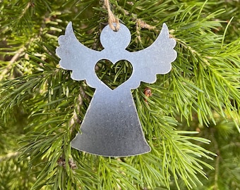 May include: A silver metal angel ornament with a heart cutout hanging on a green pine tree branch.