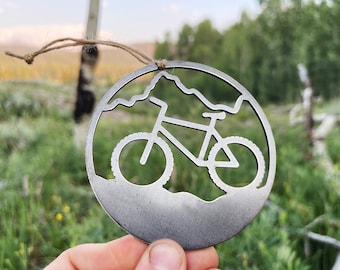 May include: A silver metal ornament shaped like a mountain range with a bicycle silhouette in the center. The ornament is hanging from a string.