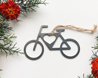 May include: A metal bicycle ornament with a heart cut out in the center. The ornament is hanging from a twine string and is surrounded by orange marigold flowers.