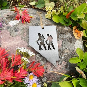 May include: A silver metal ornament shaped like the state of Nevada with two hikers cut out in the middle. The ornament is on a rock surrounded by green grass and red and white flowers.