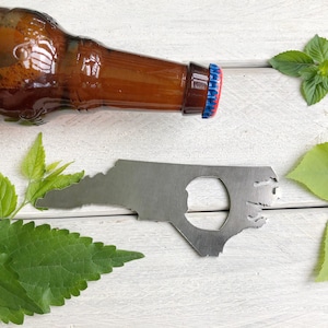 May include: A silver bottle opener shaped like the state of North Carolina. The bottle opener is on a white wooden surface with green leaves.