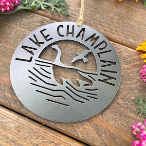 May include: A silver metal ornament with the words "Lake Champlain" cut out around the edge. The ornament features a silhouette of Champy, the lake monster, and a bird flying above the water.