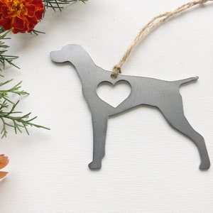 May include: A silver metal ornament shaped like a German Shorthaired Pointer dog with a heart cut out of the center. The ornament is hanging from a twine string.