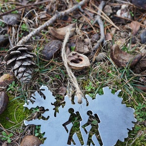 May include: A silver metal ornament shaped like the outline of Iceland with two stick figures walking inside. The ornament is hanging from a piece of twine and is lying on a bed of pine needles and moss.