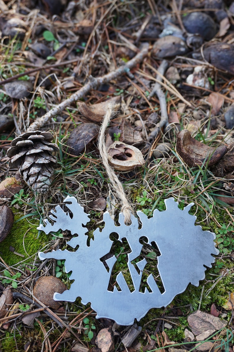 May include: A silver metal ornament shaped like the outline of Iceland with two stick figures walking inside. The ornament is hanging from a piece of twine and is lying on a bed of pine needles and moss.