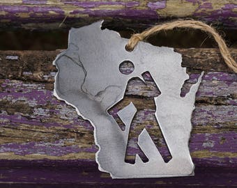 May include: A metal ornament shaped like the state of Wisconsin with a hiker silhouette inside. The ornament is hanging from a twine string and is resting on a weathered purple and brown wooden surface.
