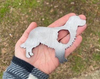 May include: A metal cutout in the shape of a dog, likely a Golden Retriever, held in a hand. The dog silhouette is detailed with a cutout in the center. The metal has a brushed finish and is silver in color.