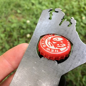 May include: A metal bottle opener shaped like the state of Michigan. The opener has a cut-out for a bottle cap and a New Belgium Brewing Company bottle cap is in the cut-out.