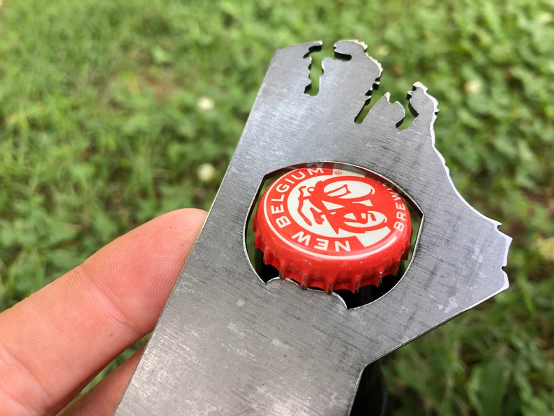 May include: A metal bottle opener shaped like the state of Michigan. The opener has a cut-out for a bottle cap and a New Belgium Brewing Company bottle cap is in the cut-out.