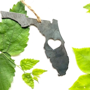 May include: A metal ornament shaped like the state of Florida with a heart cut out in the center. The ornament is a dark gray color and is surrounded by green leaves.
