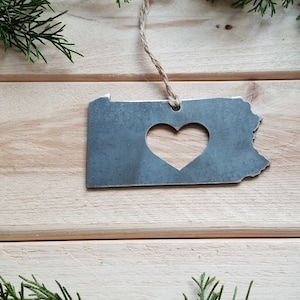May include: A metal ornament shaped like the state of Pennsylvania with a heart cut out in the center. The ornament is hanging from a twine string against a wood background.