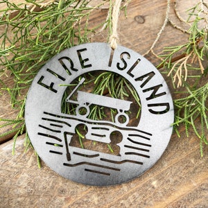 May include: A metal ornament with the words "Fire Island" and a silhouette of a wagon on a beach. The ornament is silver in color and has a rustic look.