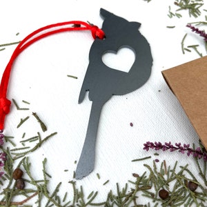 May include: A dark grey metal cardinal ornament with a heart-shaped cutout, suspended by a red cord. The bird-shaped decoration is surrounded by greenery and a small brown package.