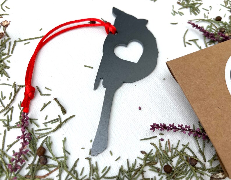 May include: A dark grey metal cardinal ornament with a heart-shaped cutout, suspended by a red cord. The bird-shaped decoration is surrounded by greenery and a small brown package.