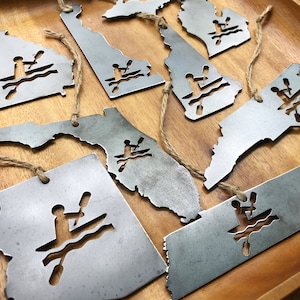 May include: A set of eight metal ornaments shaped like different states in the US. Each ornament features a silhouette of a person kayaking. The ornaments are silver in color and have a rustic finish.