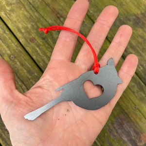 May include: A grey metal bird-shaped ornament with a heart cut-out, suspended by a red cord. The ornament is held in a hand against a wooden background.