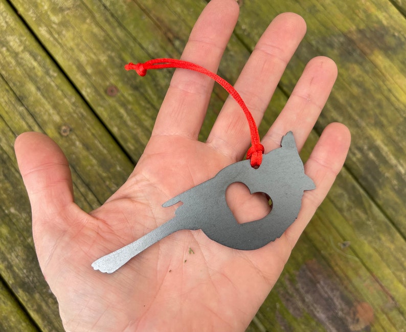 May include: A grey metal bird-shaped ornament with a heart cut-out, suspended by a red cord. The ornament is held in a hand against a wooden background.