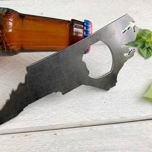 May include: A metal bottle opener shaped like the state of North Carolina. The opener is laying on a white wooden surface with a brown glass bottle and green leaves in the background.