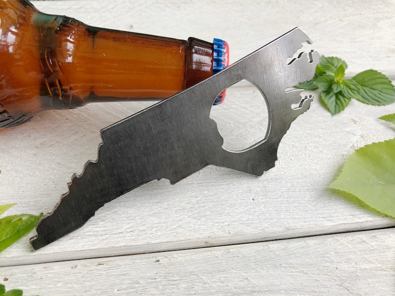 May include: A metal bottle opener shaped like the state of North Carolina. The opener is laying on a white wooden surface with a brown glass bottle and green leaves in the background.