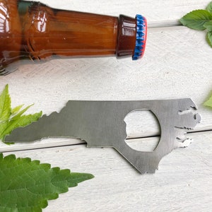 May include: A metal bottle opener shaped like the state of North Carolina. The opener has a hole in the center for opening bottles. The opener is on a white wooden surface with green leaves.