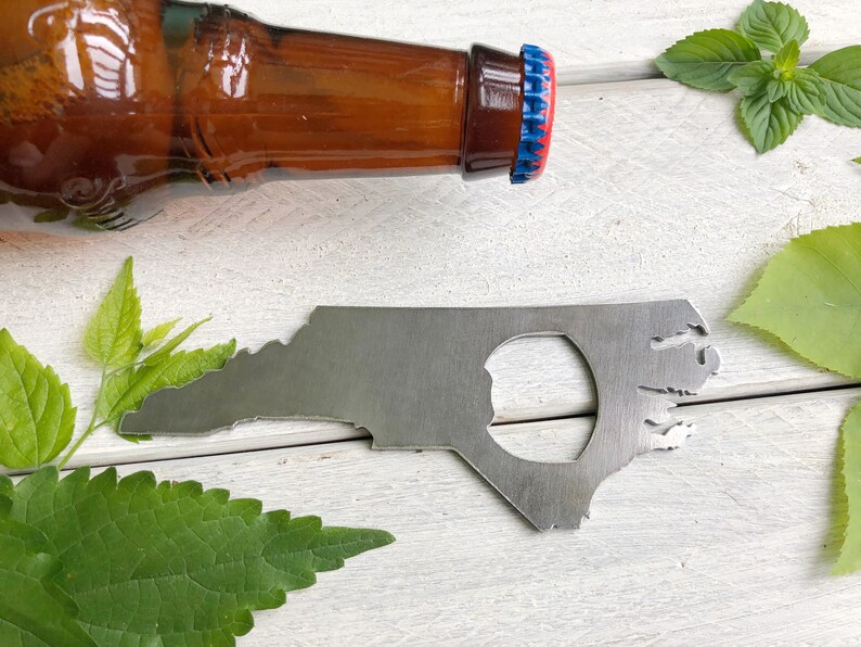 May include: A metal bottle opener shaped like the state of North Carolina. The opener has a hole in the center for opening bottles. The opener is on a white wooden surface with green leaves.