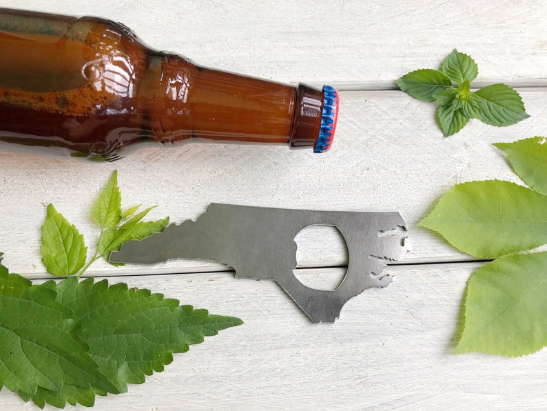 May include: A silver bottle opener shaped like the state of North Carolina. The bottle opener is on a white wooden surface with green leaves.
