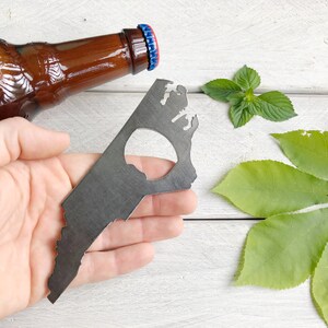 May include: A metal bottle opener shaped like the state of Virginia. The opener has a hole in the center for opening bottles. The opener is held in a hand against a white wooden background.