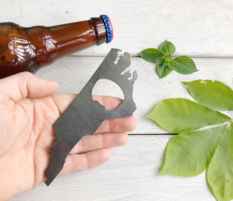 May include: A metal bottle opener shaped like the state of Virginia. The opener has a hole in the center for opening bottles. The opener is held in a hand against a white wooden background.