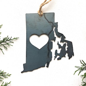May include: A metal ornament shaped like the state of Rhode Island with a heart cut out in the center. The ornament is painted a dark gray color and has a rustic finish.