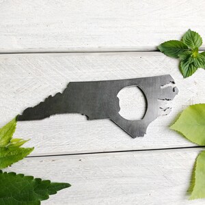 May include: A metal bottle opener shaped like the state of North Carolina. The opener has a hole in the center for opening bottles.