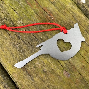 May include: A silver metal cardinal ornament with a heart-shaped cutout, suspended by a red cord. The bird-shaped decoration is displayed on a weathered wooden surface.