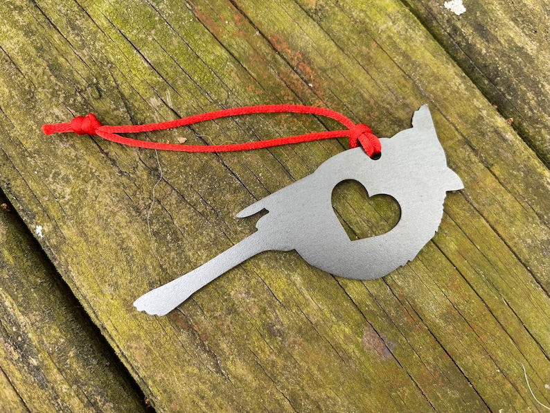 May include: A silver metal cardinal ornament with a heart-shaped cutout, suspended by a red cord. The bird-shaped decoration is displayed on a weathered wooden surface.
