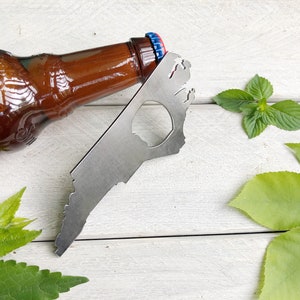 May include: A silver bottle opener shaped like the state of North Carolina. The opener is lying on a white wooden surface with green leaves around it. A brown glass beer bottle is lying on the surface next to the opener.