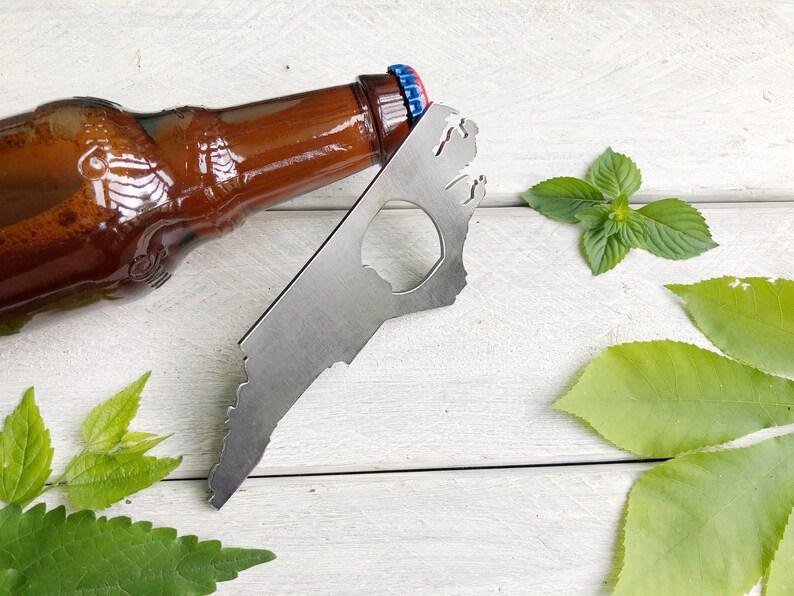 May include: A silver bottle opener shaped like the state of North Carolina. The opener is lying on a white wooden surface with green leaves around it. A brown glass beer bottle is lying on the surface next to the opener.