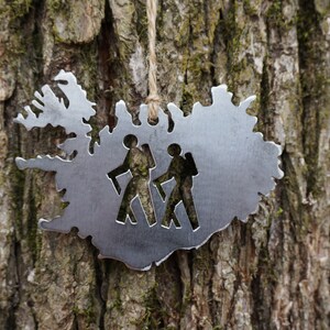 May include: A silver metal ornament shaped like the outline of Iceland with two hikers cut out inside. The ornament is hanging from a string on a tree trunk.