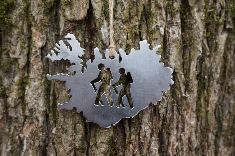 May include: A silver metal ornament shaped like the outline of Iceland with two hikers cut out inside. The ornament is hanging from a string on a tree trunk.