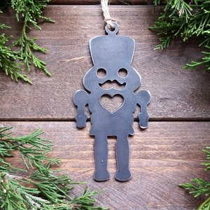 May include: A metal nutcracker ornament with a heart-shaped cutout on the chest. The ornament is painted black and has a rustic finish. It is hanging from a twine string.