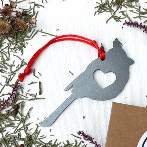 May include: A grey metal cardinal ornament with a heart-shaped cutout. The bird-shaped decoration has a red string for hanging. The ornament is surrounded by greenery and small brown seed pods.