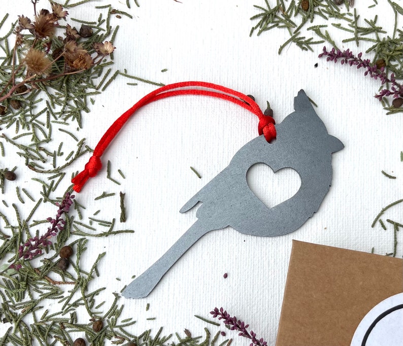 May include: A grey metal cardinal ornament with a heart-shaped cutout. The bird-shaped decoration has a red string for hanging. The ornament is surrounded by greenery and small brown seed pods.
