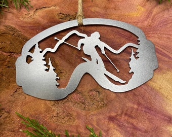 Puede incluir: Un adorno de metal plateado con forma de gafas de esquí con una silueta de un esquiador en las montañas en su interior. El adorno tiene una cuerda de yute para colgar.