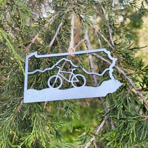 May include: A metal ornament shaped like the state of Pennsylvania with a silhouette of a bicycle inside. The ornament is hanging from a tree branch.
