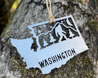 May include: A metal ornament shaped like the state of Washington with a silhouette of Bigfoot, trees, and stars. The ornament is hanging from a brown string and has the word "WASHINGTON" written on it.