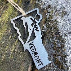 May include: A silver metal ornament shaped like the state of Vermont with a skier silhouette and the word "VERMONT" inscribed on it. The ornament is hanging from a twine string.